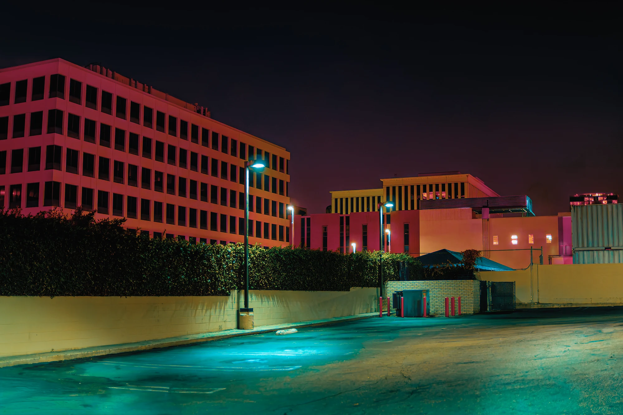 Pink-lit office buildings behind a hedge, an empty parking lot glowing cyan