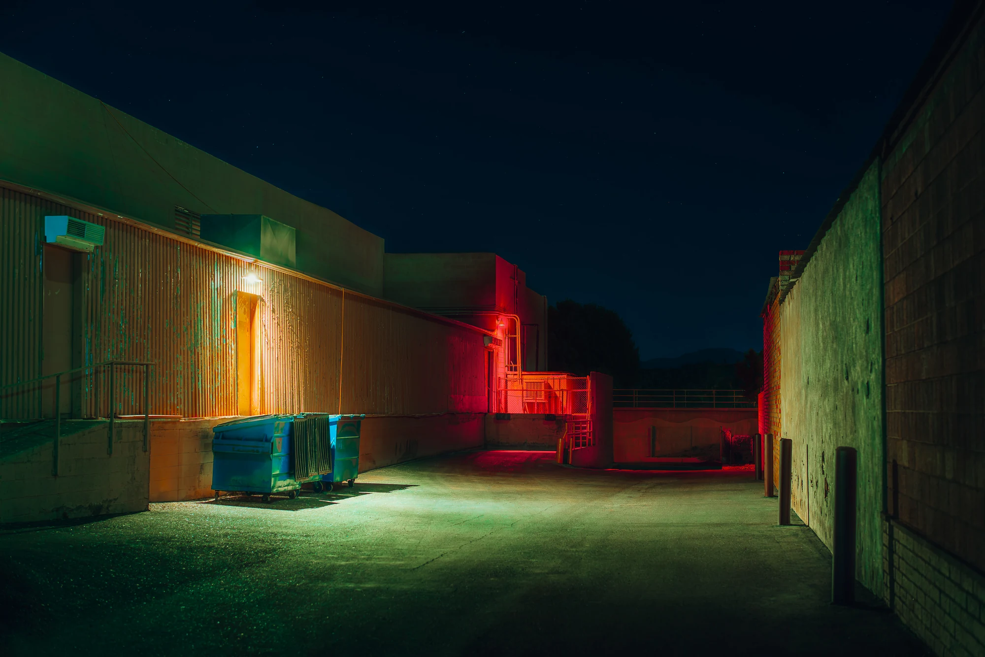Empty service alley at night, blue dumpsters under green light and a staircase glowing red at the far end