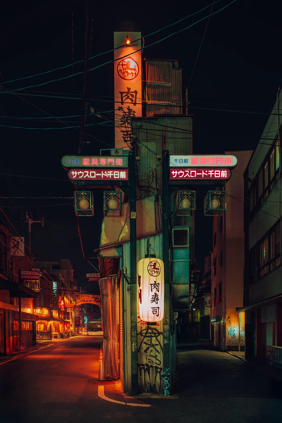 Street corner with a tall pole of signs, a lit paper lantern and neon arches in Japanese, power lines overhead at night.