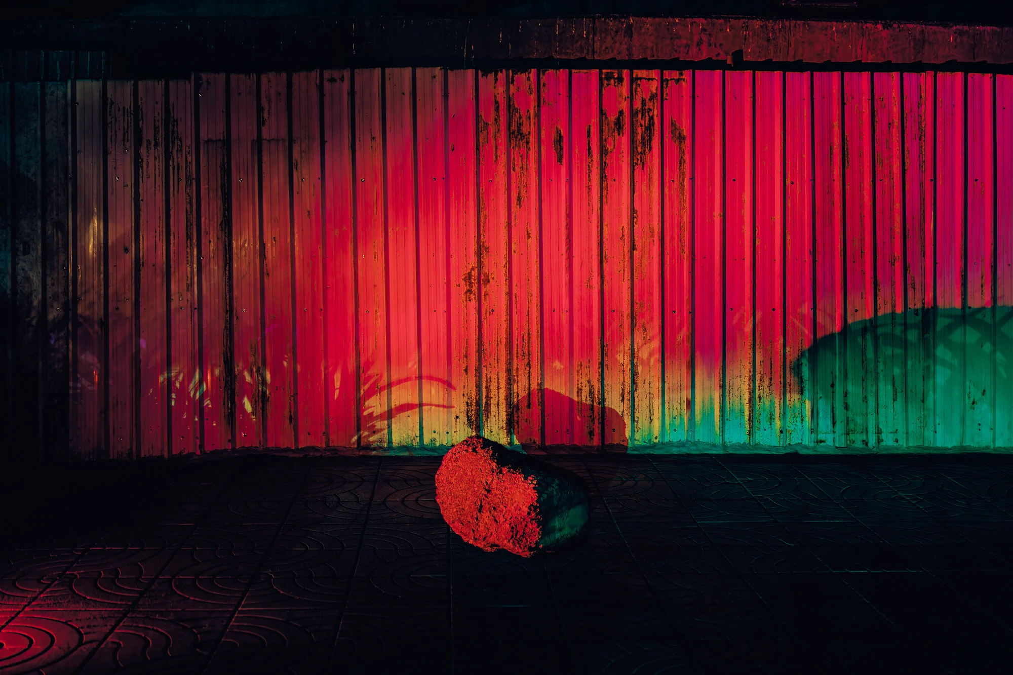 A lone stone in front of a corrugated metal fence lit red and green