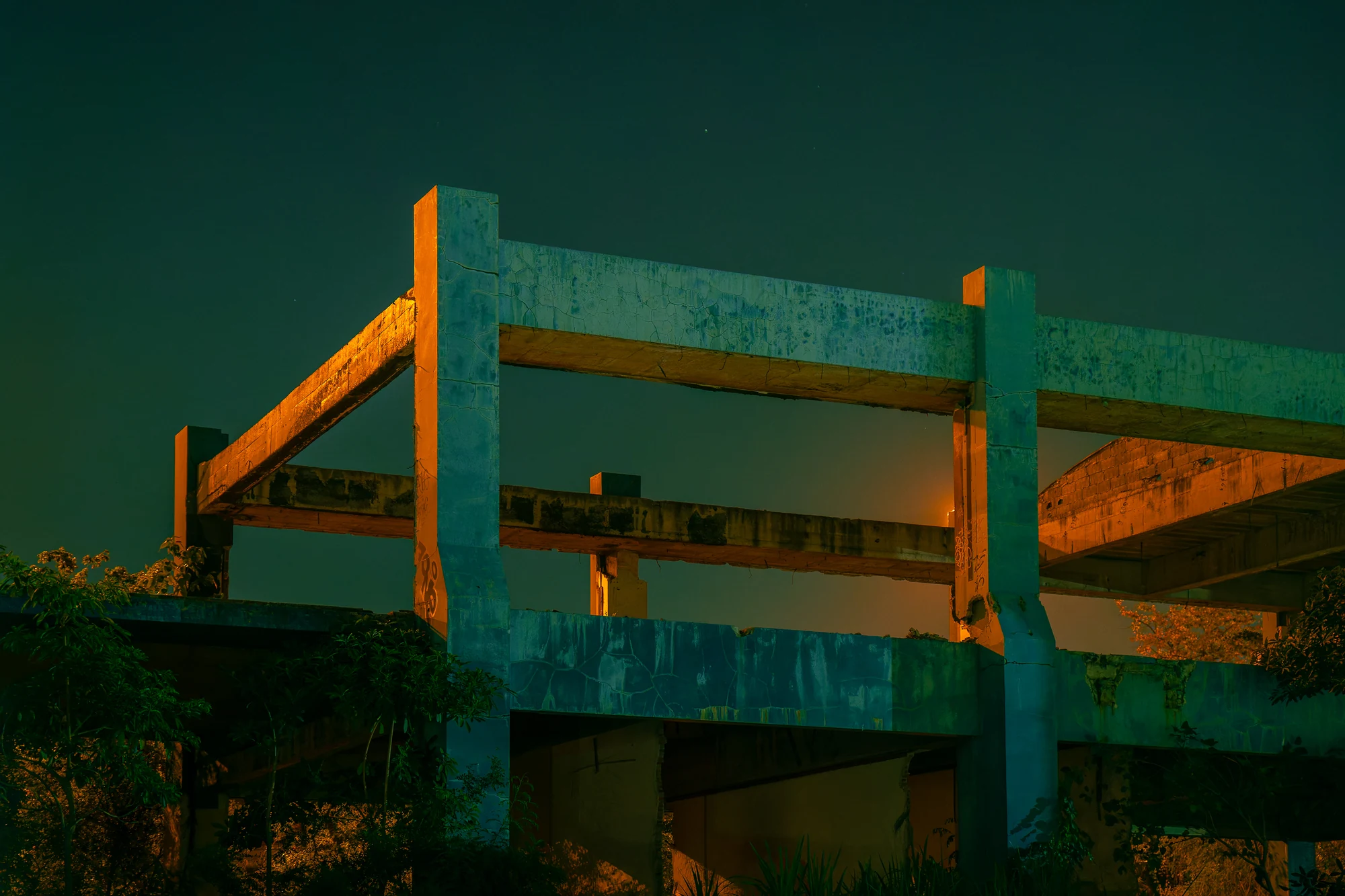 The bare concrete frame of an abandoned building lit teal and orange above trees